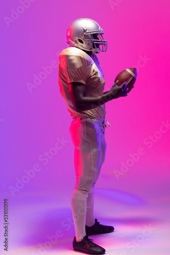 African american male american football player holding ball with neon purple and pink lighting