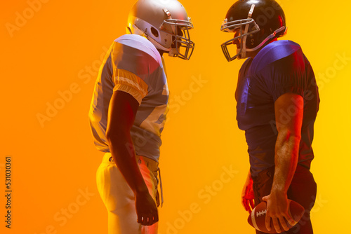 Diverse male american football players holding ball with neon orange lighting