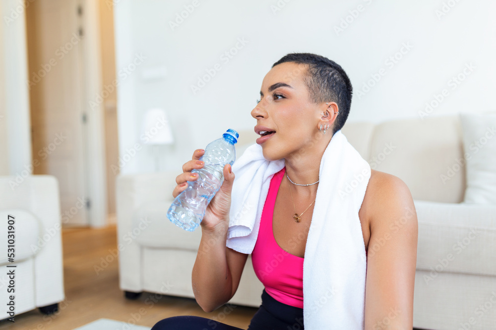 Young adult African American woman drinking water from plastic bottle, sitting on fitness mat and resting after training at home Young adult African American woman drinking water from plastic bottle, sitting on fitness mat and resting after training at home
