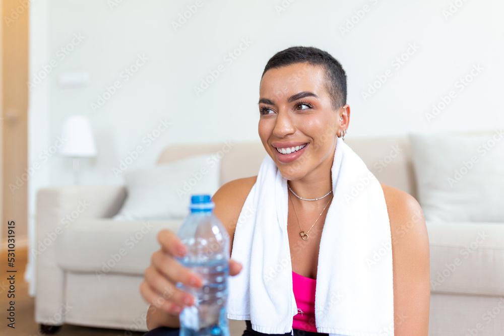 Young adult African American woman drinking water from plastic bottle, sitting on fitness mat and resting after training at home Young adult African American woman drinking water from plastic bottle, sitting on fitness mat and resting after training at home