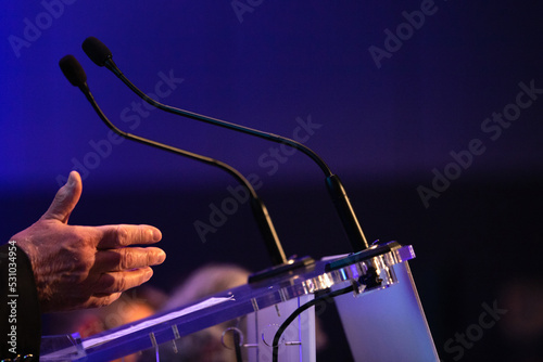 focus sur un micro et les mains d'un orateur prononçant un discours à la tribune lors d'un meeting politique, d'un colloque ou d'une réunion	 photo