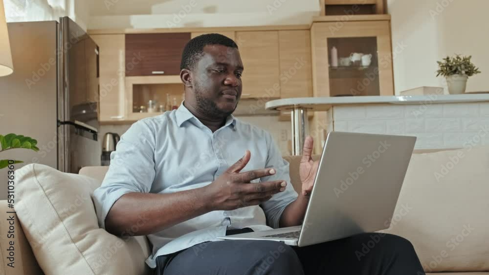 Medium shot of young Black man in light blue office shirt video conferencing on laptop while sitting on couch in living room, working from home Medium shot of young Black man in light blue office shirt video conferencing on laptop while sitting on couch in living room, working from home