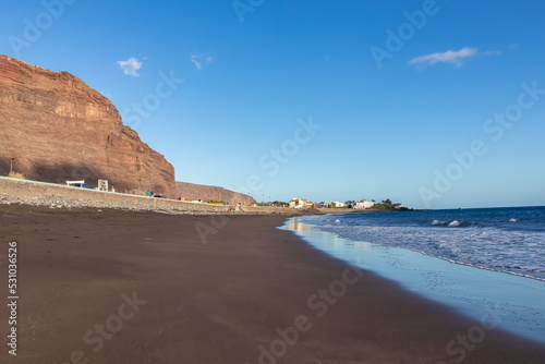 Scenic view on beach Playa Valle Gran Rey at early morning seen from Promenade La Calera in Valle Gran Rey on La Gomera, Canary Islands, Spain, Europe. Mountain reflections in dark volcanic sand beach