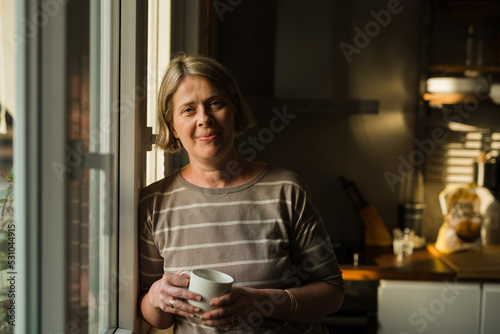 mid adult woman standing by the kitchen window and drinking coffee