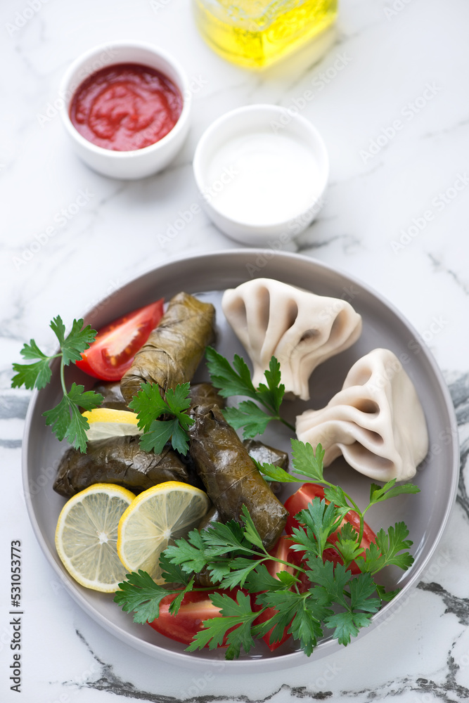 Plate with dolma or stuffed grape leaves, khinkali and dips on a white marble background, vertical shot, selective focus Plate with dolma or stuffed grape leaves, khinkali and dips on a white marble background, vertical shot, selective focus