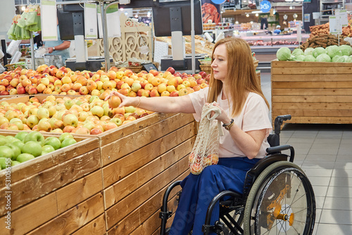 Woman shopper who uses a wheelchair ,chooses apples in a supermarket, puts them in mesh bag © evafesenuk