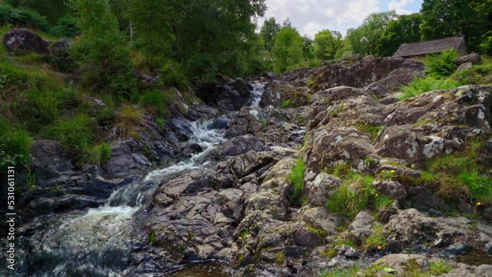 Slow moving woodland stream with ferns on the riverbank and water flowing over the rocks. Shot in Cumbria the Lake District England. Slow moving woodland stream with ferns on the riverbank and water flowing over the rocks. Shot in Cumbria the Lake District England.