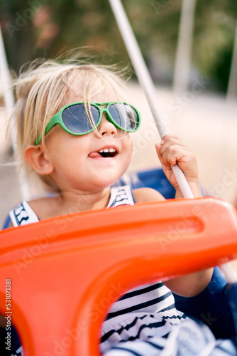 Little girl in sunglasses swings on a swing with her tongue out for joy. High quality photo