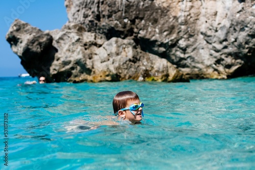 Child boy with swimming goggles swims in the sea next to a rock during a happy summer vacation in Corfu, Greece