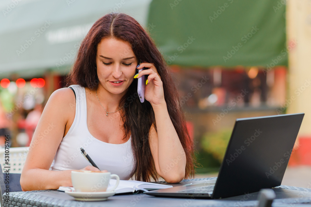 Attractive woman having a call and using her laptop while sitting outdoors at the cafe terrace.Summer nice evening.