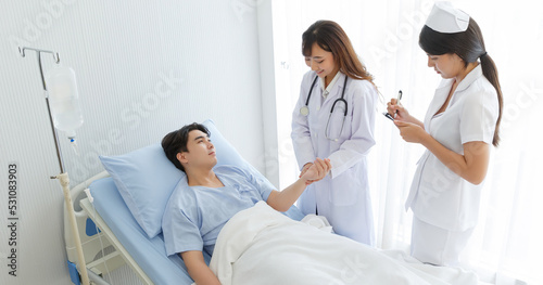 Female doctor and young male patient who lie on the bed while checking pulse, consult and explain with nurse taking note and supporting in hospital wards.