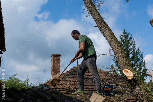 A man cleans the roof of a house after a big storm on a summer day. A man stands on the roof and removes fallen leaves and branches