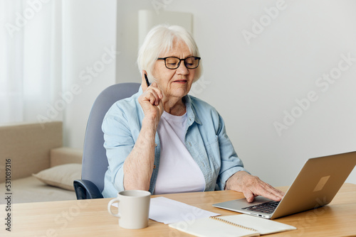 an attentive, focused elderly businesswoman is sitting at home in a bright interior at work at a laptop and, while conducting video training, looks attentively, holding her index finger near her face