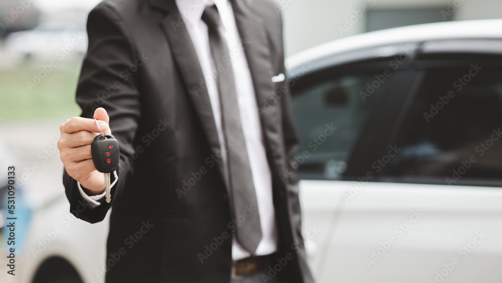 A man in a suit stands holding the car keys next to a white car, getting a new car with a car showroom dealer. Car trading concept.