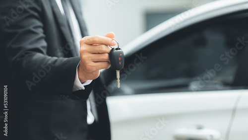 A man in a suit stands holding the car keys next to a white car, getting a new car with a car showroom dealer. Car trading concept.