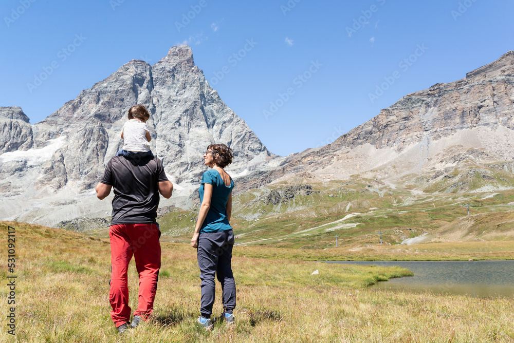 Standing father carrying his little daughter on shoulders and his smiling wife during a hiking day in Cervinia, italian alps. 