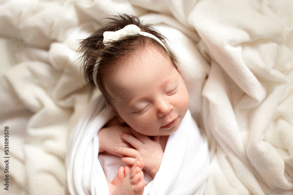 Top view of a newborn baby girl sleeping in a white cocoon on a white bed. Beautiful portrait of a little girl 7 days, one week. Macro studio professional photography. Top view of a newborn baby girl sleeping in a white cocoon on a white bed. Beautiful portrait of a little girl 7 days, one week. Macro studio professional photography.