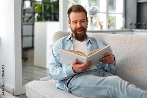 Young bearded handsome man reading book on couch at home