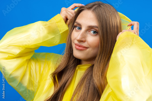 Young blonde woman wearing yellow raincoat smiling at camera over blue background in studio photo