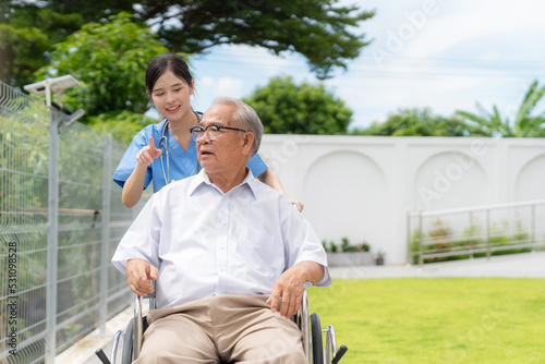 Young Asia caregiver doctor pushing elderly man in wheelchair in the garden to get refreshment at nursing home. Mental care and wellness. Nurse taking care old man and physical therapy
