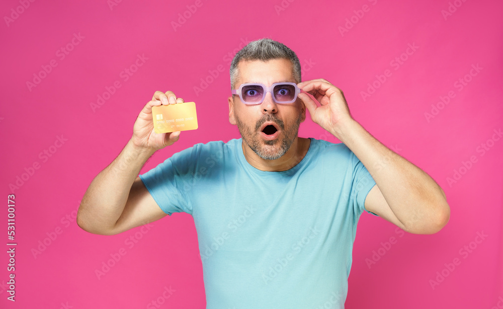 Excited of a new offer handsome man hold debit, credit card in hand lowering his blue glasses wearing blue t-shirt isolated on pink background. Man with bank card in hand. Financial, banking concept