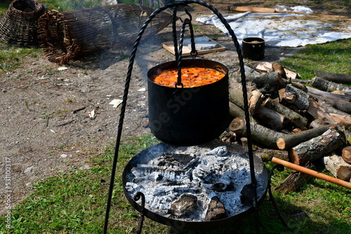 A close up on a pot with goulash or stew boiling after being cooked on a temporary metal grill with charred and burning logs still beneath the pot seen on a sunny summer day in Poland