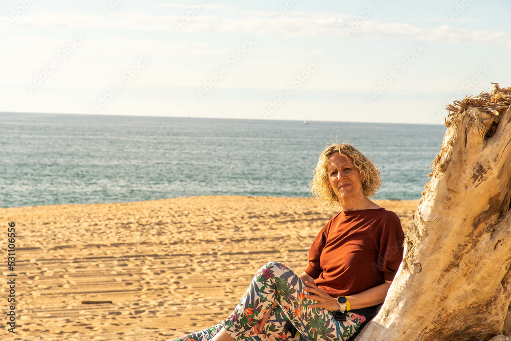 blonde beautiful woman leaning against a tree trunk on the beach