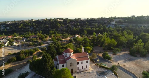 Aerial drone footage of Archangel Michael church in Souni Zanakia, Limassol, Cyprus. 360 view of bell tower, square, sea, Souni Zanatzia forest and ceramic tile roof architecture at sunset from above