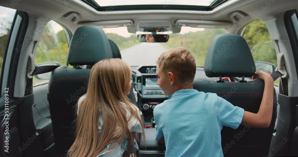 Back view. Kids looking outside and pretty woman riding by road wheel. Child boy and girl cheerful parent spending time together in car.