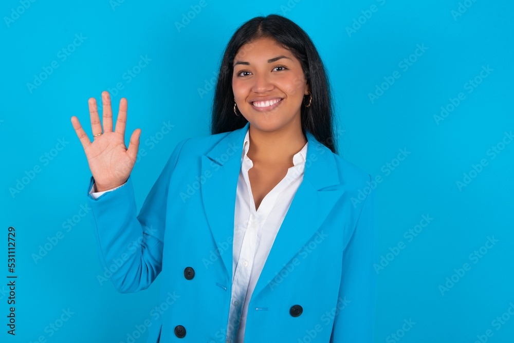 Young latin woman wearing blue blazer blue background Waiving saying hello happy and smiling, friendly welcome gesture. Young latin woman wearing blue blazer blue background Waiving saying hello happy and smiling, friendly welcome gesture.