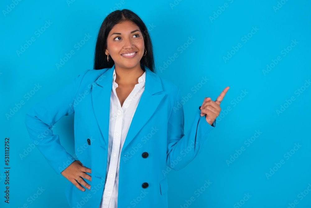 Smiling Young latin woman wearing blue blazer blue background indicating finger empty space showing best low prices Smiling Young latin woman wearing blue blazer blue background indicating finger empty space showing best low prices