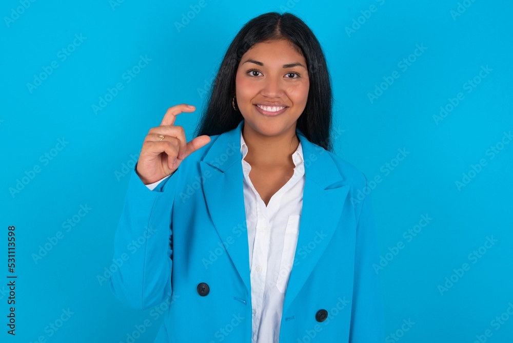 Young latin woman wearing blue blazer blue background smiling and confident gesturing with hand doing small size sign with fingers looking and the camera. Measure concept Young latin woman wearing blue blazer blue background smiling and confident gesturing with hand doing small size sign with fingers looking and the camera. Measure concept