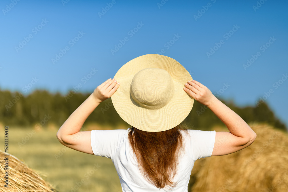 Back view of woman, girl in straw hat standing on yellow farm meadow with ripe golden wheat and against the blue sky. Harvesting concept. Vacation concept