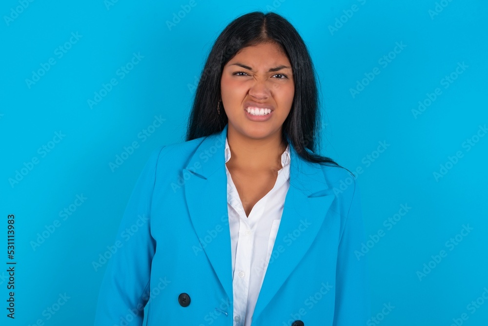 Young latin woman wearing blue blazer blue background keeps teeth clenched, frowns face in dissatisfaction, irritated because of much duties. Young latin woman wearing blue blazer blue background keeps teeth clenched, frowns face in dissatisfaction, irritated because of much duties.