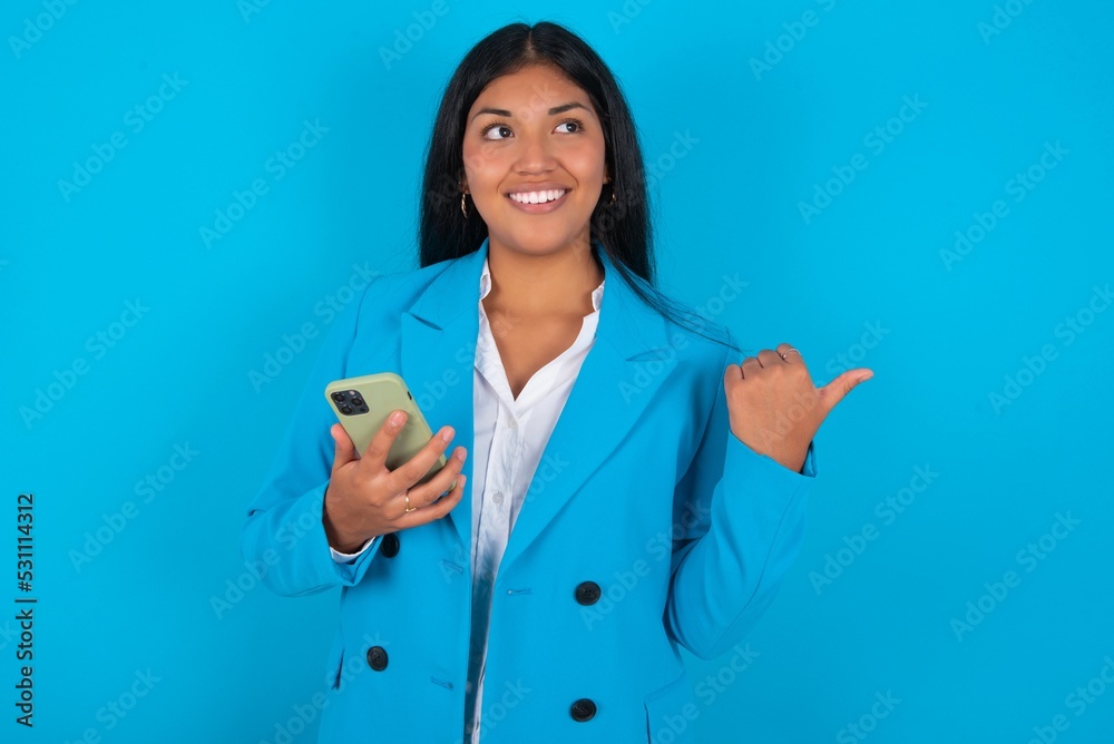 Young latin woman wearing blue blazer blue background using and texting with smartphone pointing and showing with thumb up to the side with happy face smiling Young latin woman wearing blue blazer blue background using and texting with smartphone pointing and showing with thumb up to the side with happy face smiling