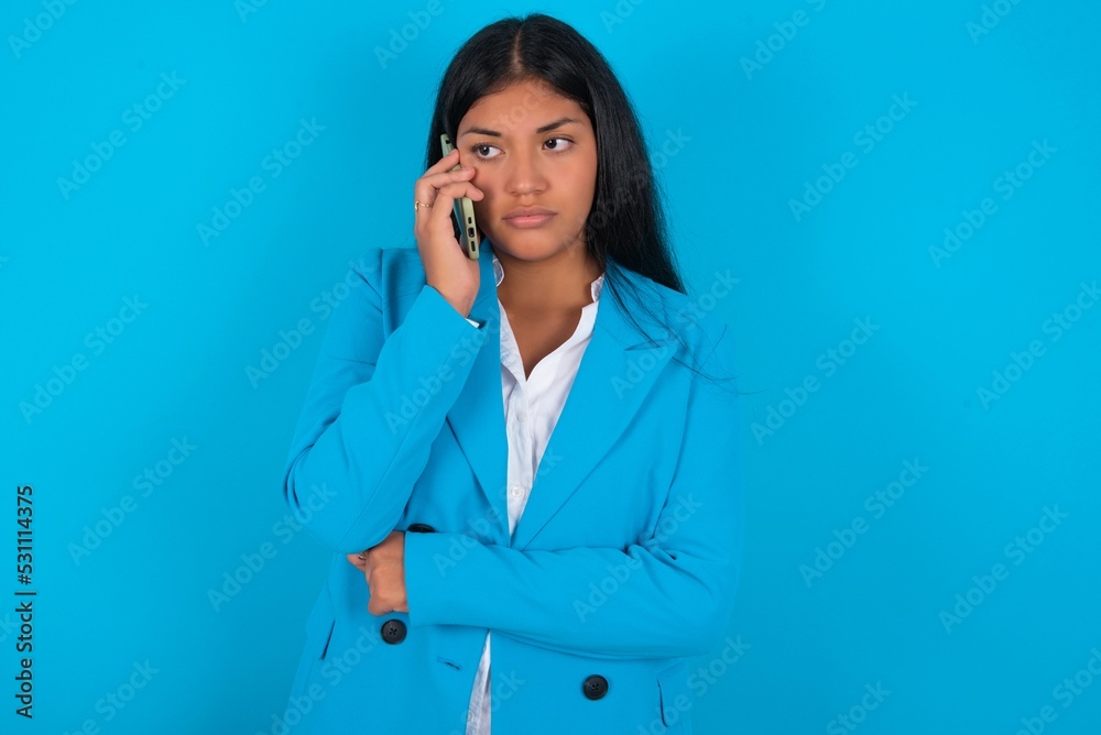 Sad Young latin woman wearing blue blazer blue background talking on smartphone. Communication concept. Sad Young latin woman wearing blue blazer blue background talking on smartphone. Communication concept.