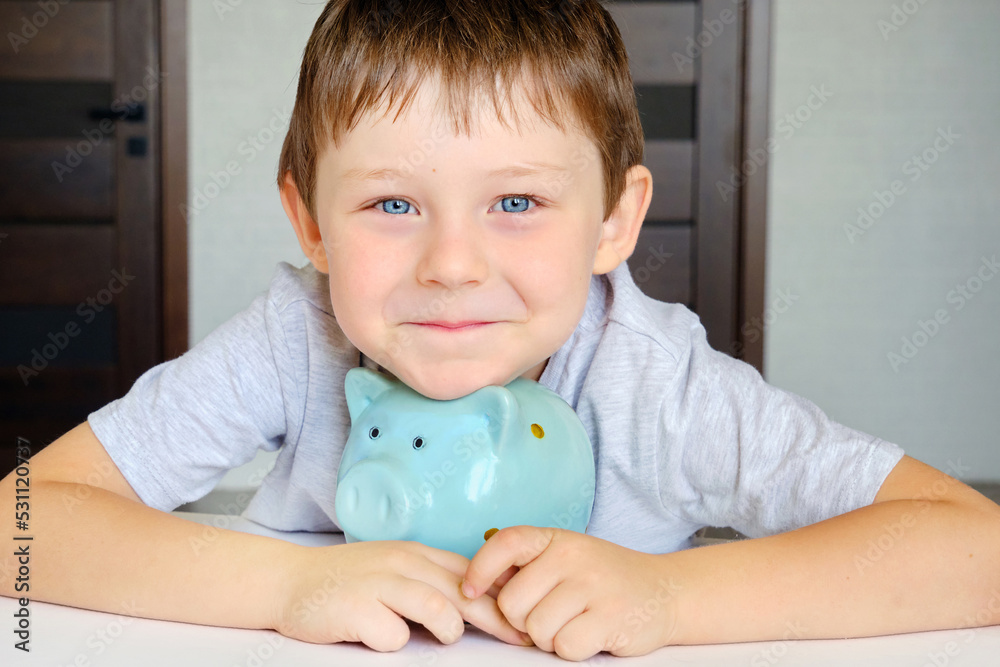 a cute blue-eyed boy with a smile on his face hugs his piggy bank, a blue pig. The child looks at the camera, horizontal photo a cute blue-eyed boy with a smile on his face hugs his piggy bank, a blue pig. The child looks at the camera, horizontal photo