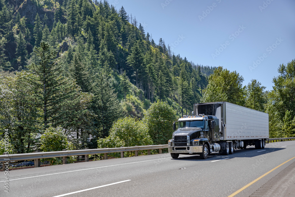 Industrial big rig black semi truck tractor transporting frozen cargo in refrigerator semi trailer driving on the highway road with mountain on the side.