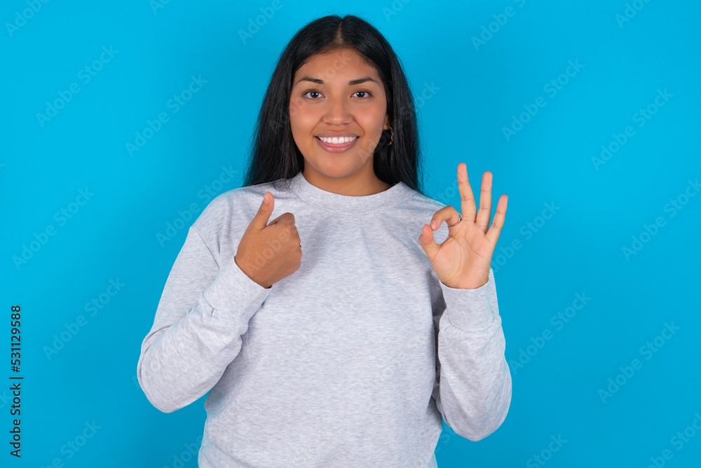 Young latin woman wearing gray sweater blue background feeling happy, amazed, satisfied and surprised, showing okay and thumbs up gestures, smiling Young latin woman wearing gray sweater blue background feeling happy, amazed, satisfied and surprised, showing okay and thumbs up gestures, smiling