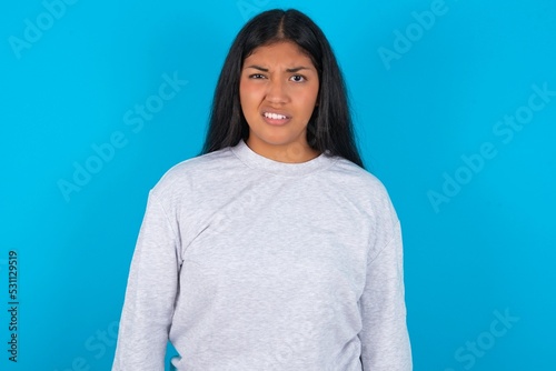 Portrait of dissatisfied Young latin woman wearing gray sweater blue background smirks face, purses lips and looks with annoyance at camera, discontent hearing something unpleasant