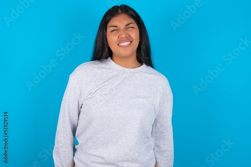 Positive Young latin woman wearing gray sweater blue background with overjoyed expression closes eyes and laughs shows white perfect teeth