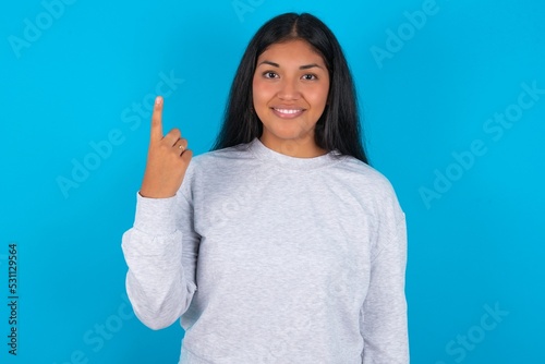 Young latin woman wearing gray sweater blue background smiling and looking friendly, showing number one or first with hand forward, counting down