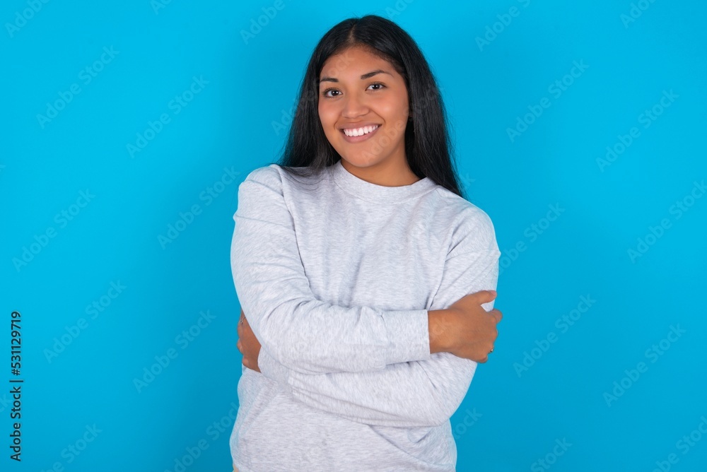 Charming pleased Young latin woman wearing gray sweater blue background embraces own body, pleasantly feels comfortable poses. Tenderness and self esteem concept Charming pleased Young latin woman wearing gray sweater blue background embraces own body, pleasantly feels comfortable poses. Tenderness and self esteem concept