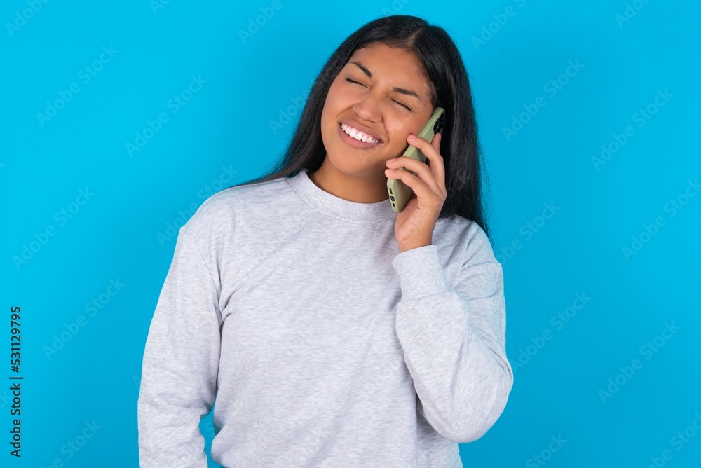 Funny Young latin woman wearing gray sweater blue background laughs happily, has phone conversation, being amused by friend, closes eyes. Funny Young latin woman wearing gray sweater blue background laughs happily, has phone conversation, being amused by friend, closes eyes.