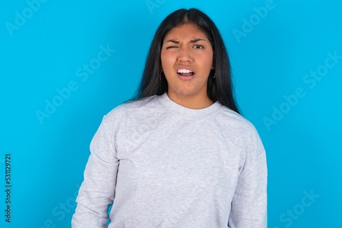 Young latin woman wearing gray sweater blue background yawns with opened mouth stands. Daily morning routine