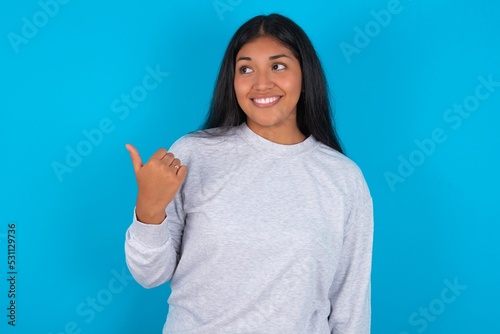Young latin woman wearing gray sweater blue background points away and gives advice demonstrates advertisement