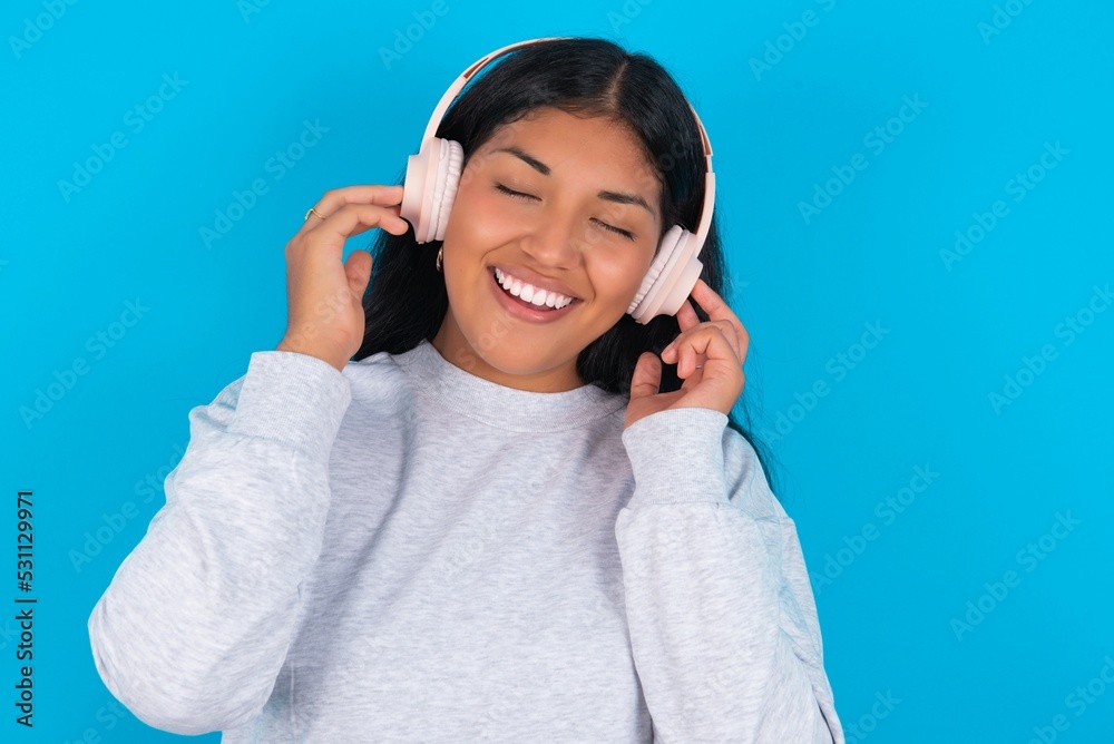 Young latin woman wearing gray sweater blue background smiles broadly feels very glad listens favourite music track via wireless headphones closes eyes. Young latin woman wearing gray sweater blue background smiles broadly feels very glad listens favourite music track via wireless headphones closes eyes.