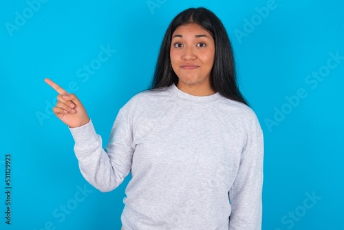 Positive Young latin woman wearing gray sweater blue background with satisfied expression indicates at upper right corner shows good offer suggests to click on link