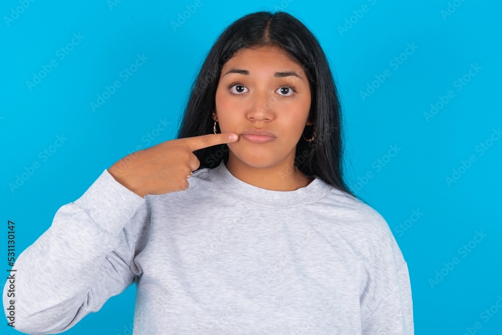 Charming Young hispanic woman wearing grey sweatshirt over blue background pointing on pout lips with forefinger, showing effect after lifting procedure, Charming Young hispanic woman wearing grey sweatshirt over blue background pointing on pout lips with forefinger, showing effect after lifting procedure,