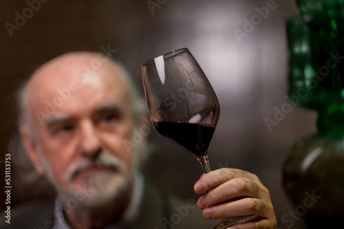 Senior graybeard Caucasian man, a wine connoisseur, holding a glass, analyzing the red wine through sight, smell, and taste photo